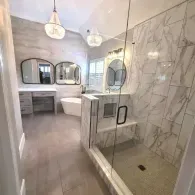 A modern bathroom featuring a walk-in glass shower, marble-tiled walls, a double vanity with two mirrors, and a tub.