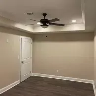 An empty room with beige walls, dark wood floors, a white door, and a ceiling fan in a recessed tray ceiling.