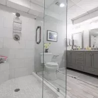 A modern, bright bathroom with a glass-enclosed shower, a white toilet, and a grey vanity with a mirror.