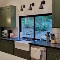 A modern kitchen features olive-green cabinets, black countertops, a white farmhouse sink, and three wall-mounted lamps.