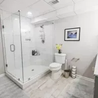 Modern bathroom with a glass shower enclosure, white toilet, light wood-look floors, and framed wall art.