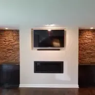 A wall with a recessed TV above a fireplace, flanked by dark cabinets and textured stone accents on either side.