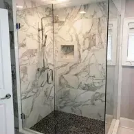 A walk-in shower with marble-patterned tile walls, glass doors, and a dark pebble floor.