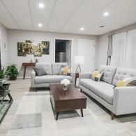 A bright, modern living room with light gray sofas, a dark wood coffee table, and a multi-panel art piece on the wall.