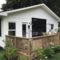 A white building with horizontal siding features a deck with a wooden railing and a glass garage door beside a side door.