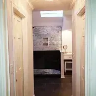 A view through a hallway into a modern bathroom with dark flooring, stone-tiled walls, and a small vanity area.