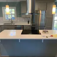 A modern kitchen island with a white countertop and black cooktop, featuring a stainless steel vent hood and pendant lights.