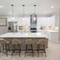 A modern kitchen with a white quartz island, four beige bar stools, white cabinetry, and stainless steel appliances.