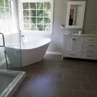 A modern bathroom featuring a white freestanding tub, gray walls, a window with white trim, and a white vanity with mirror.