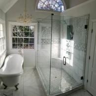 Modern bathroom with a white claw-foot tub, a glass-enclosed shower, a chandelier, and an arched window.