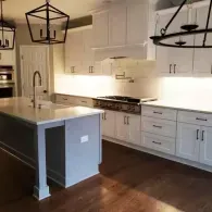 A bright, modern kitchen with white cabinets, a gray island, dark wood floors, and two large hanging geometric lanterns.