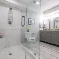 A modern, white-tiled bathroom featuring a glass shower stall, a white toilet, and a grey vanity with a mirror.