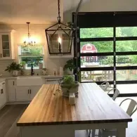 A kitchen with a wood-topped island, a farmhouse sink, and a modern lantern light, opening to a view of a red barn.