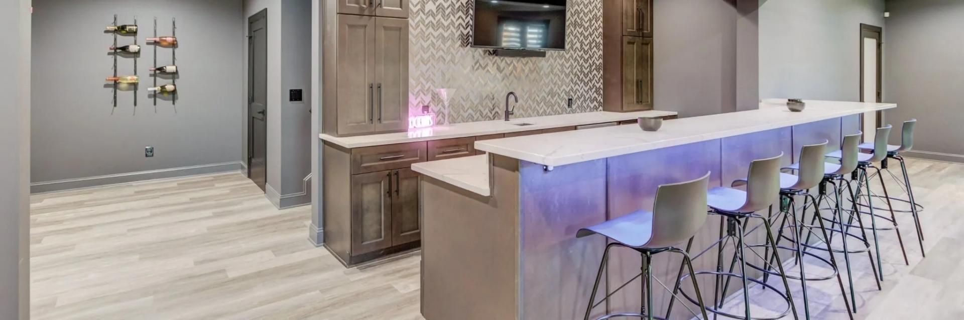 A modern basement wet bar with grey walls, light wood floors, a long white countertop, and six bar stools with purple light.