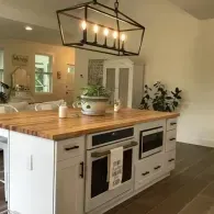 A white kitchen island with a butcher block countertop, featuring a built-in oven, microwave, and a black metal chandelier.