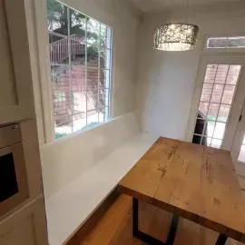 A dining nook with a white built-in bench, a wooden table, and a decorative pendant light next to a large window and door.