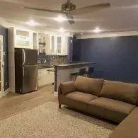 A modern living room with a beige sofa, a neutral area rug, a breakfast bar, and a stainless steel refrigerator.