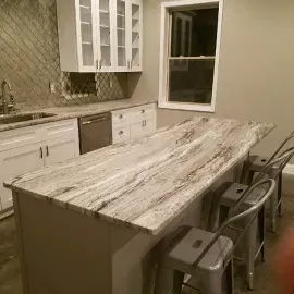 A modern kitchen island with a white and gray marbled countertop and three gray metal bar stools, in front of cabinetry.