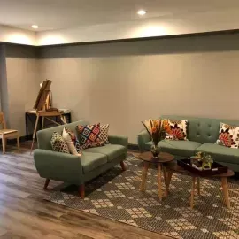 A living room features two sage-green sofas, a patterned area rug, two wooden coffee tables, and an artist's easel.