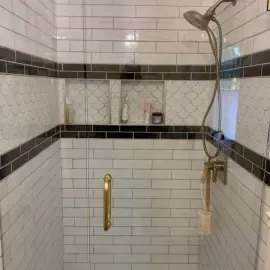 A tiled shower with white subway tiles, a black accent stripe, scallop-patterned niches, and gold hardware.
