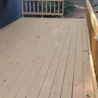 A light brown wooden deck with matching railing, viewed from above, leading toward a dirt path in a yard.