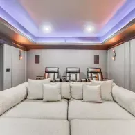 A home theater with a large white lounge sofa in the foreground facing three leather recliners under ambient lighting.