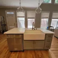 A kitchen island with a stainless steel dishwasher and white farmhouse sink, featuring gray cabinets and wood flooring.