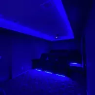A home theater room with tiered seating illuminated by vibrant blue LED accent lighting along the floor and ceiling.