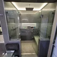 A modern shower enclosure with gray tiled walls, a glass door, a stone bench, and an overhead rain shower head.