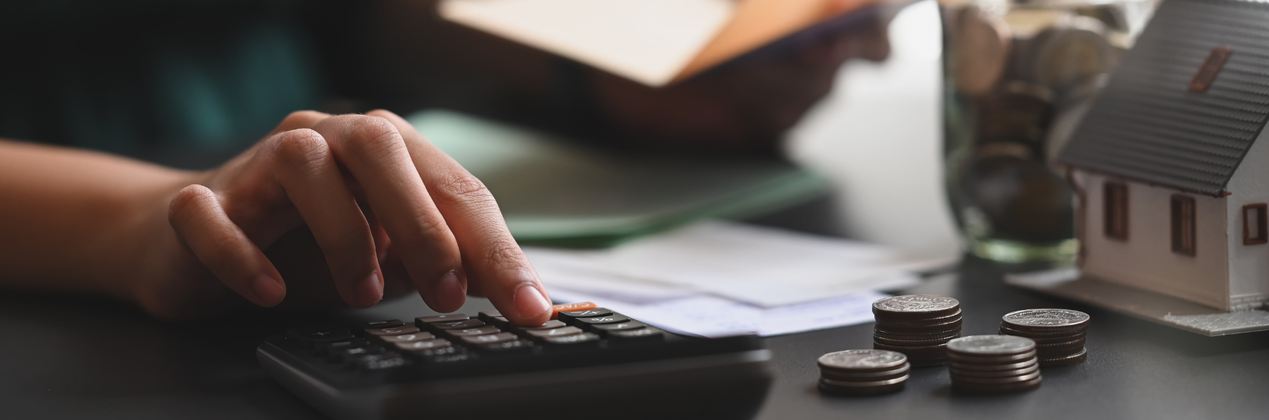 A person uses a calculator next to stacks of coins and a small model house, suggesting financial planning for a home.