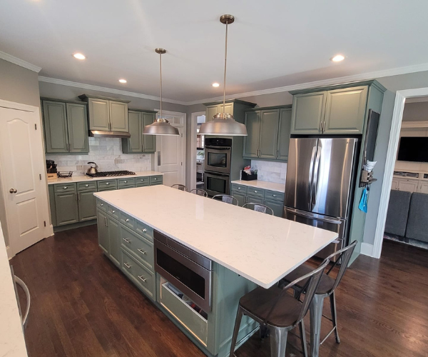 A kitchen with sage green cabinets, a large white kitchen island, stainless steel appliances, and dark wood flooring.