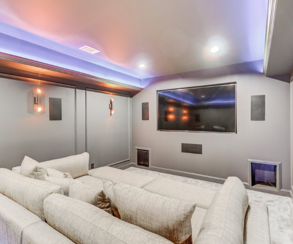 A modern home theater with a large wall-mounted TV, recessed purple lighting, and a spacious, light-colored sectional sofa.