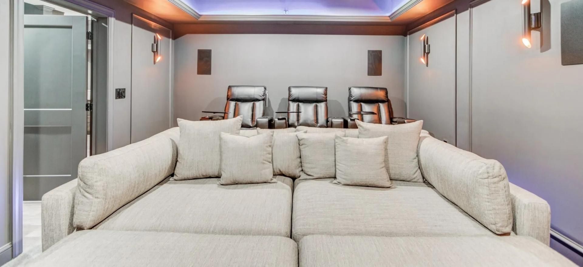 A home theater room with beige modular sofa seating in the foreground and three leather recliners against the back wall.