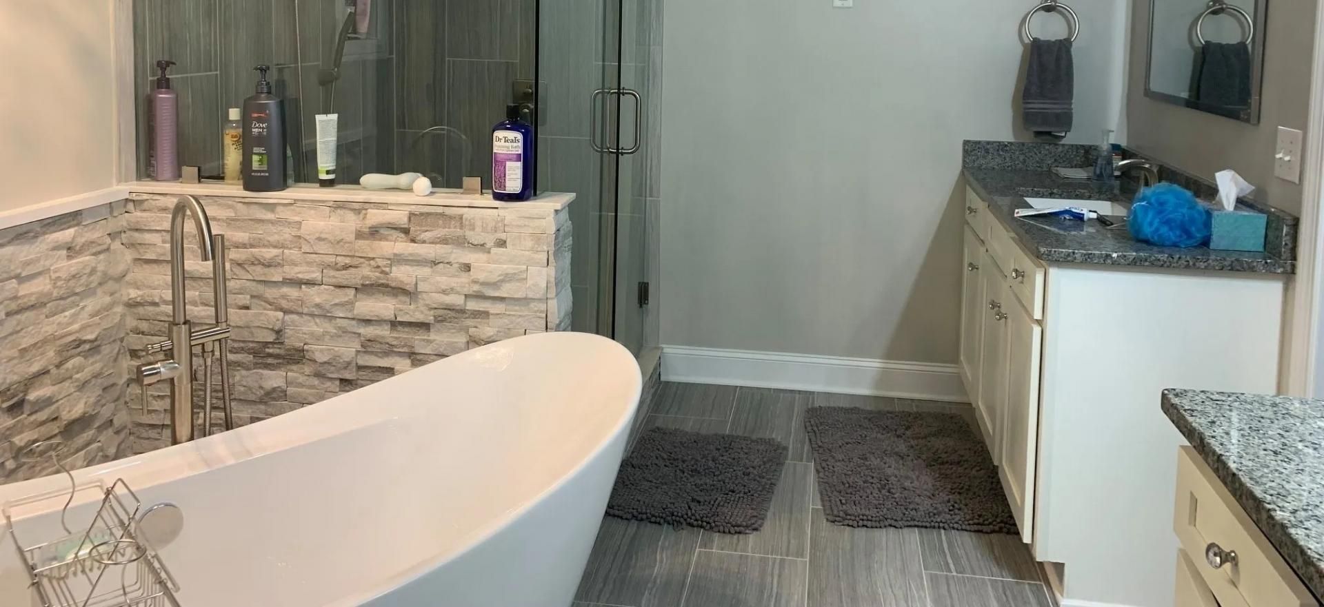 Modern bathroom featuring a white freestanding tub against a stone wall, grey tile flooring, and a vanity with a mirror.