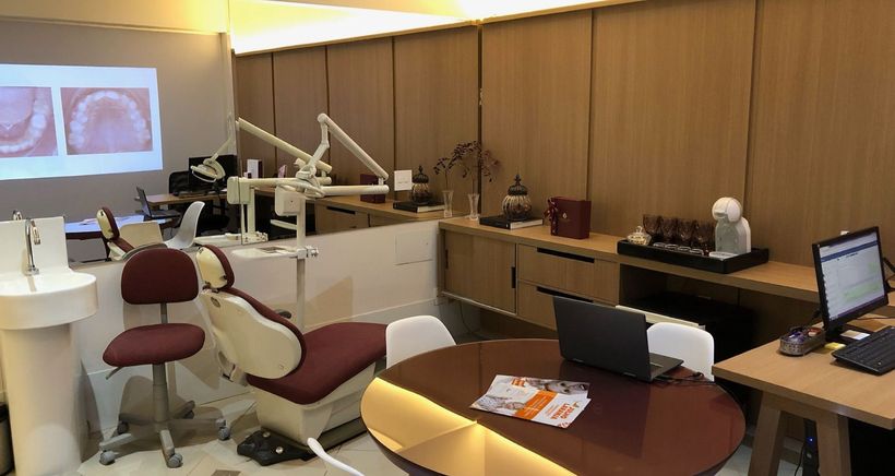 Consultório odontológico com mesa, cadeiras, computadores e tela de projeção.