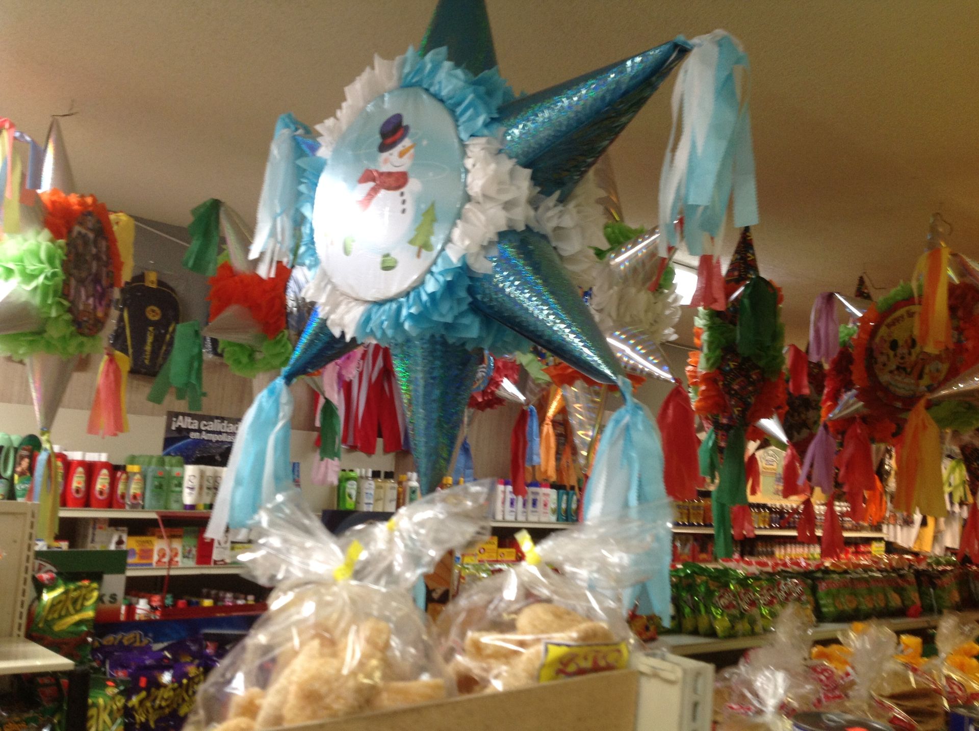 Blue star-shaped piñata with snowman design hangs above wrapped treats in a store, other piñatas in background.