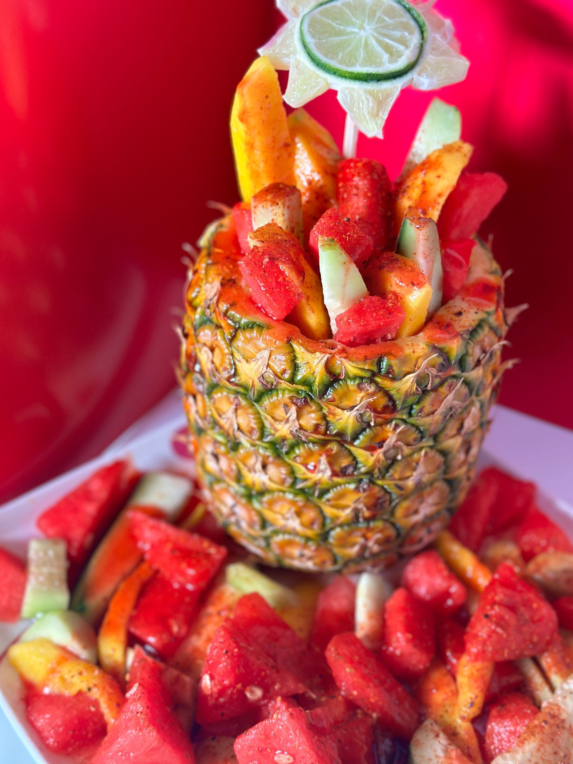 Pineapple fruit bowl filled with watermelon, mango, and cucumber; on a white plate, red background.