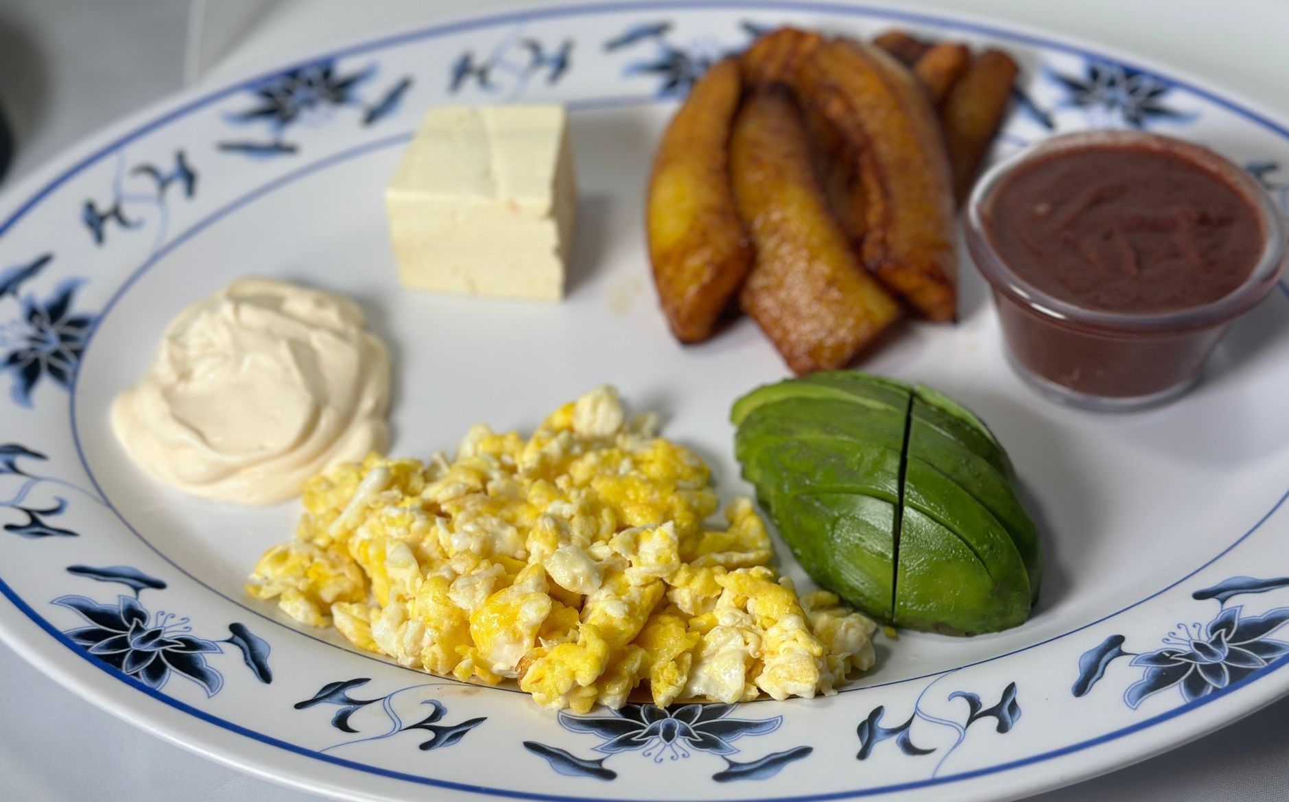 Plate with scrambled eggs, fried plantains, avocado, beans, cheese, and cream.