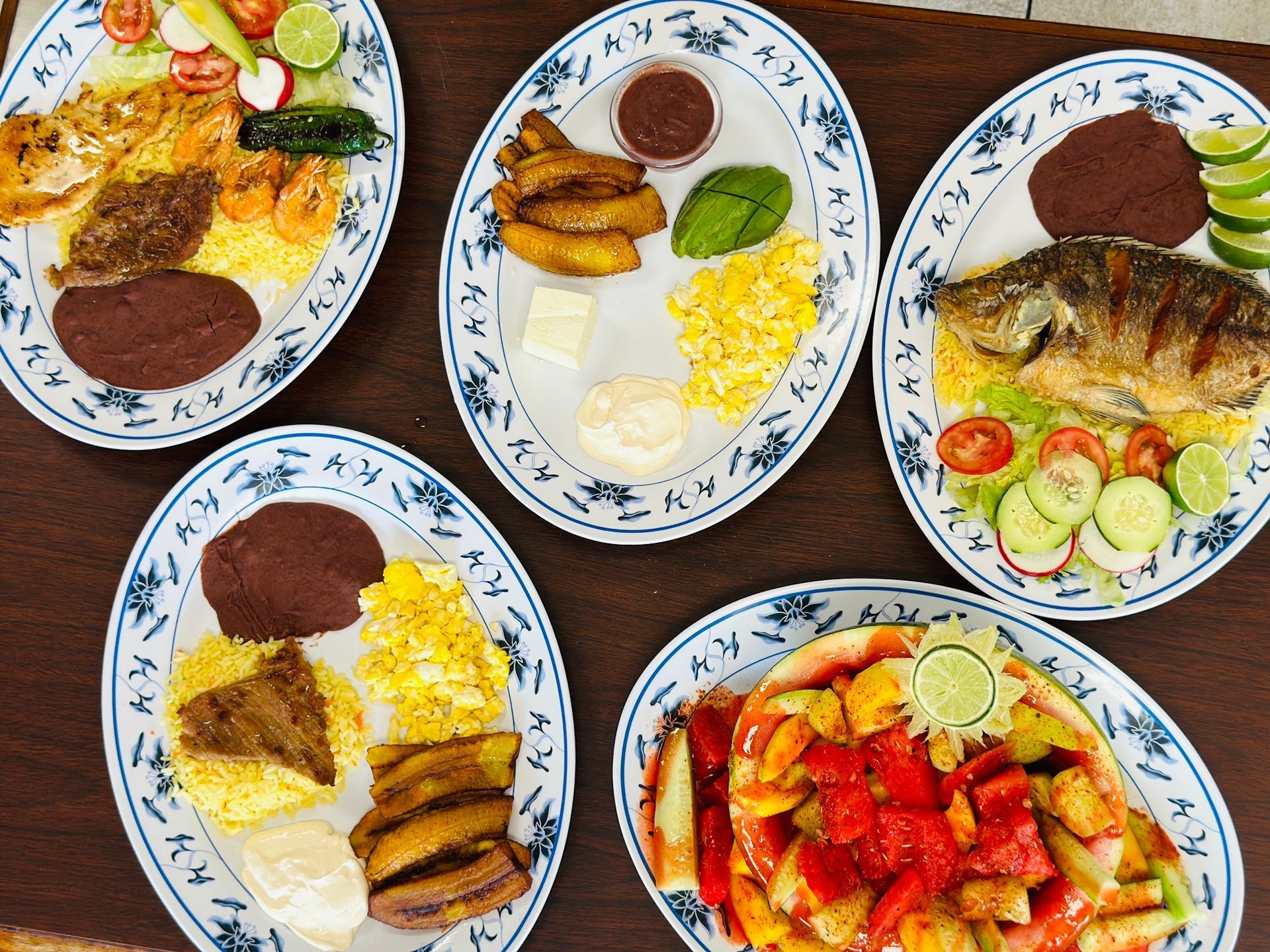 Plates of food: grilled fish, shrimp, plantains, beans, corn, vegetables. Overhead shot on a wooden table.