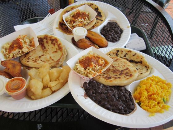 Plates of Salvadoran food: pupusas, fried plantains, beans, yellow rice, and sauces.