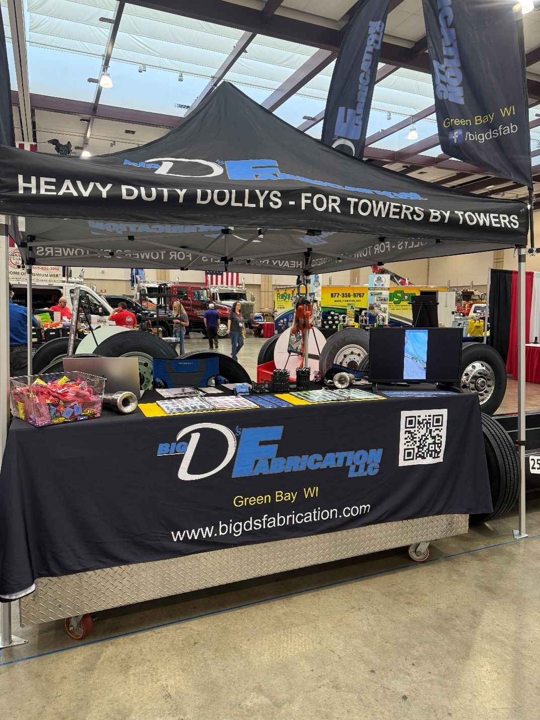 A booth at a trade show displaying heavy-duty dolly products. The booth has black and blue branding and is staffed by a person. A booth at a trade show displaying heavy-duty dolly products. The booth has black and blue branding and is staffed by a person.