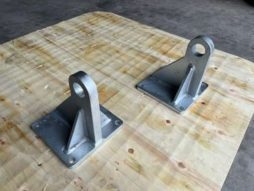 Two metal brackets with eyelets sit on a wood surface. They have a gray, industrial appearance.