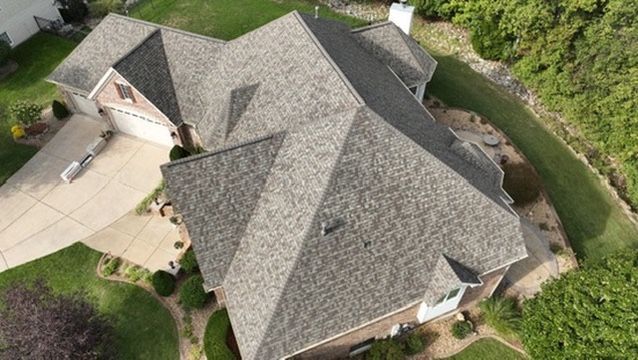 An aerial view of a single-family house with a gray shingled roof, a paved driveway, and surrounding green landscaping.