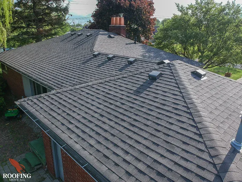 An elevated view of a residential grey shingle roof with a brick chimney and several roof vents against a treed background.