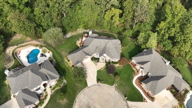 Aerial view of three large residential homes with grey roofs, surrounding driveways, a swimming pool, and wooded yards.