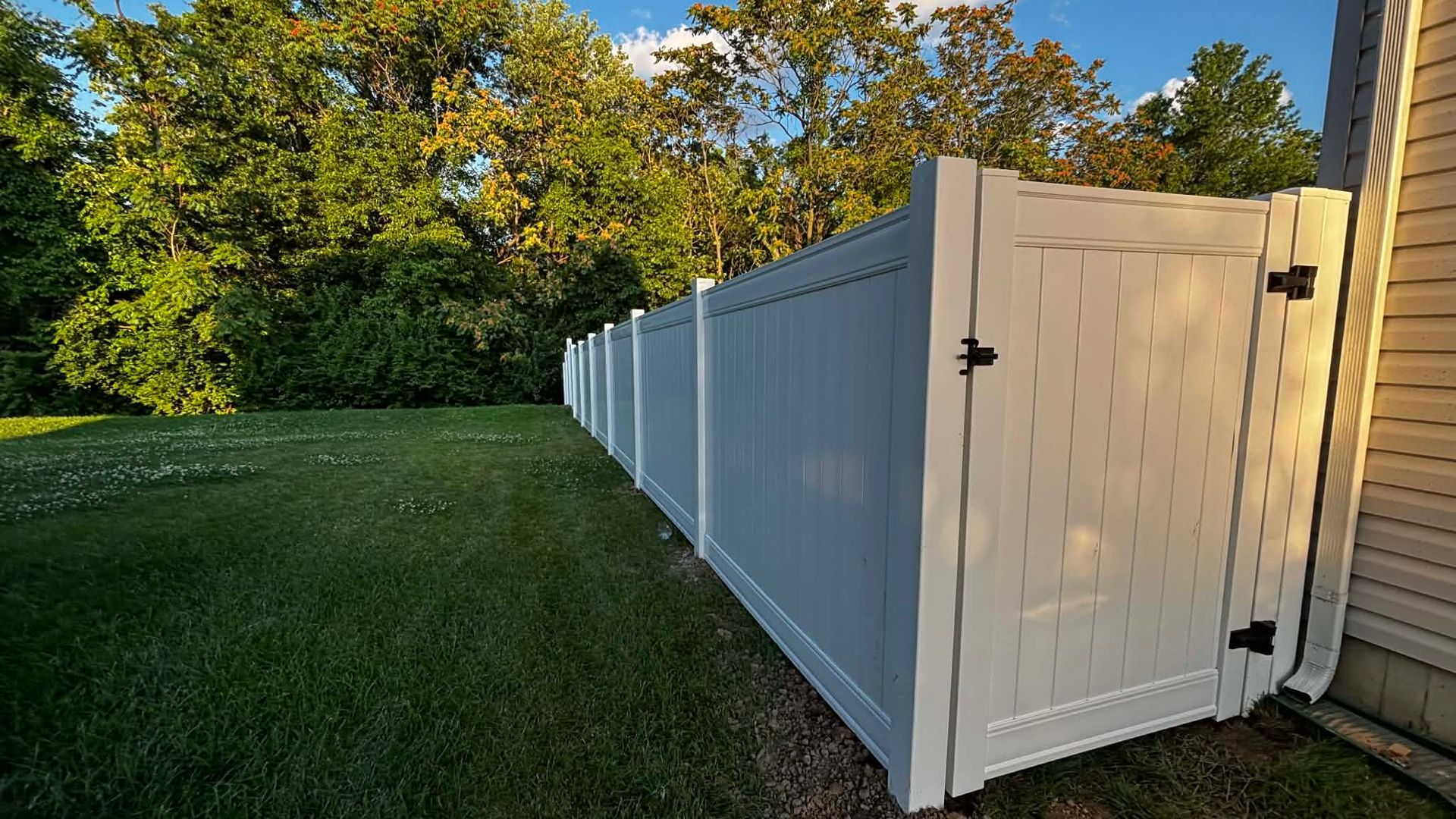 A white vinyl privacy fence runs alongside the side of a house, bordering a green lawn with a line of trees in the back.