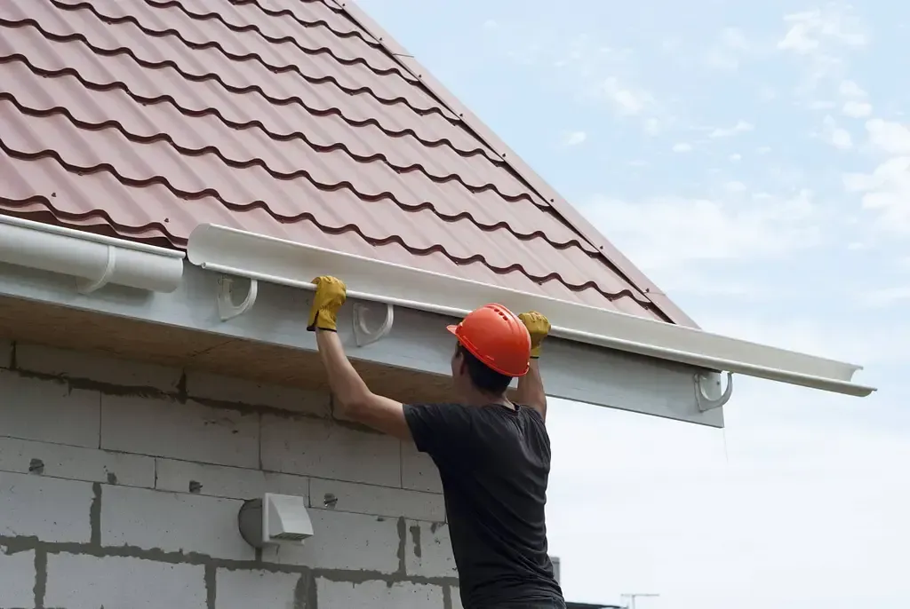 A person wearing a hard hat and gloves installs white gutters onto the edge of a roof against a brick wall.