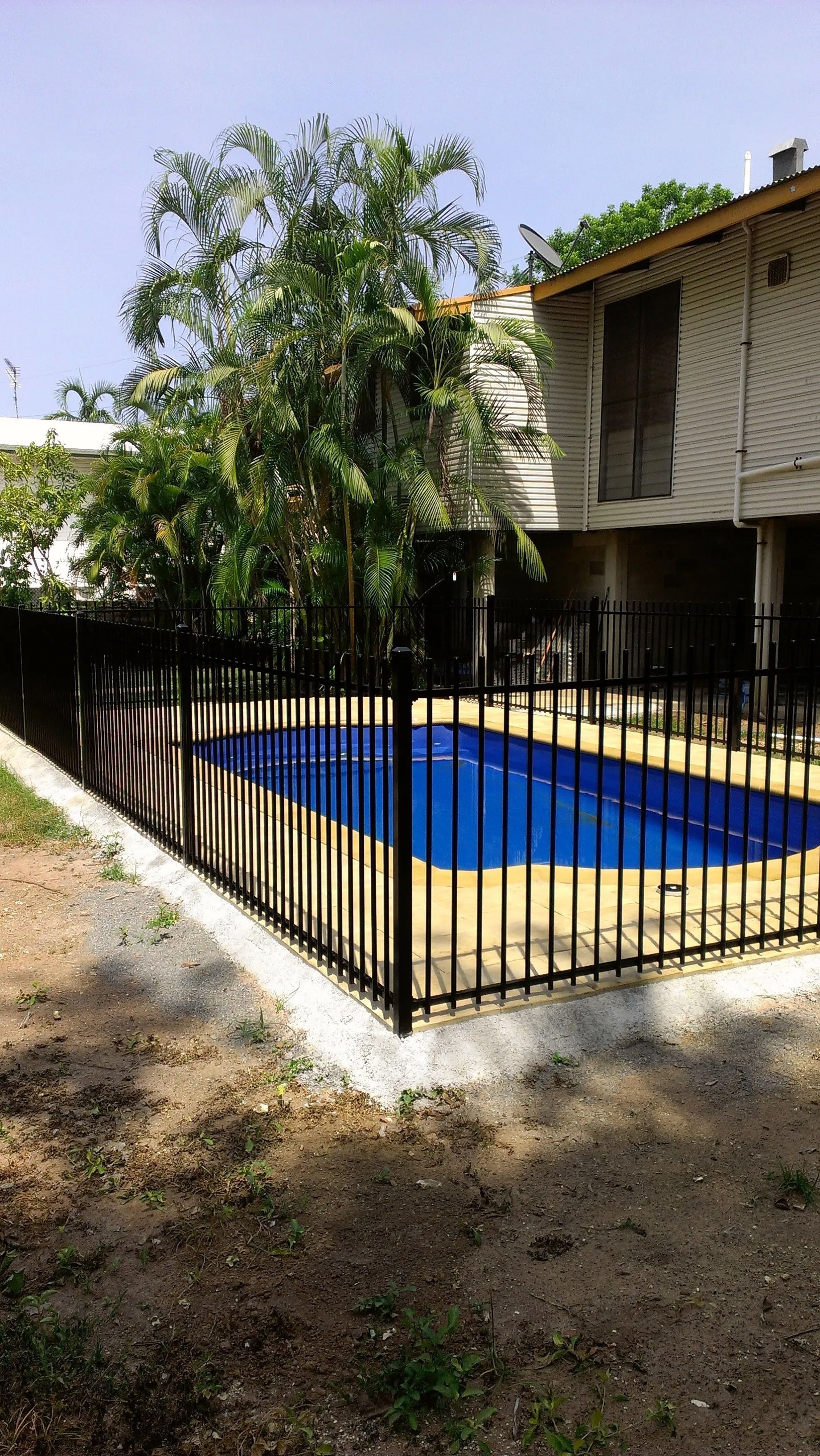 Black fenced blue swimming pool next to a light-coloured two-story building and trees. — Fence Factory in Berrimah, NT