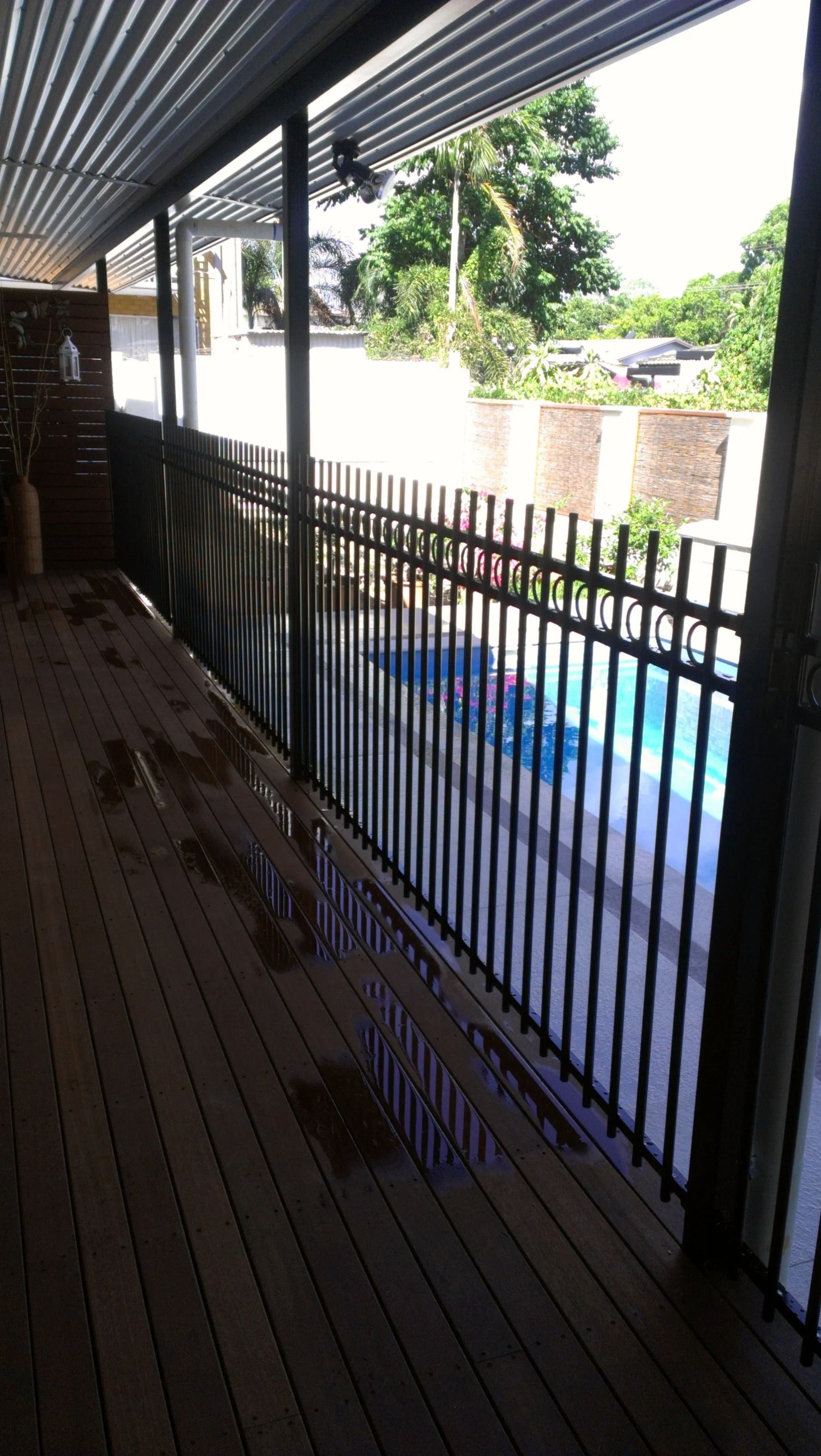 Wooden deck with black fence overlooking a swimming pool. Trees and a building are in the background. — Fence Factory in Berrimah, NT