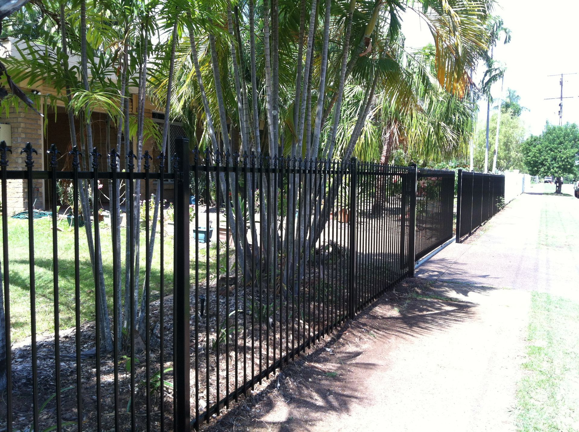 A Black Fence is Surrounded by Grass and Trees in a Backyard — Fence Factory In Berrimah, NT